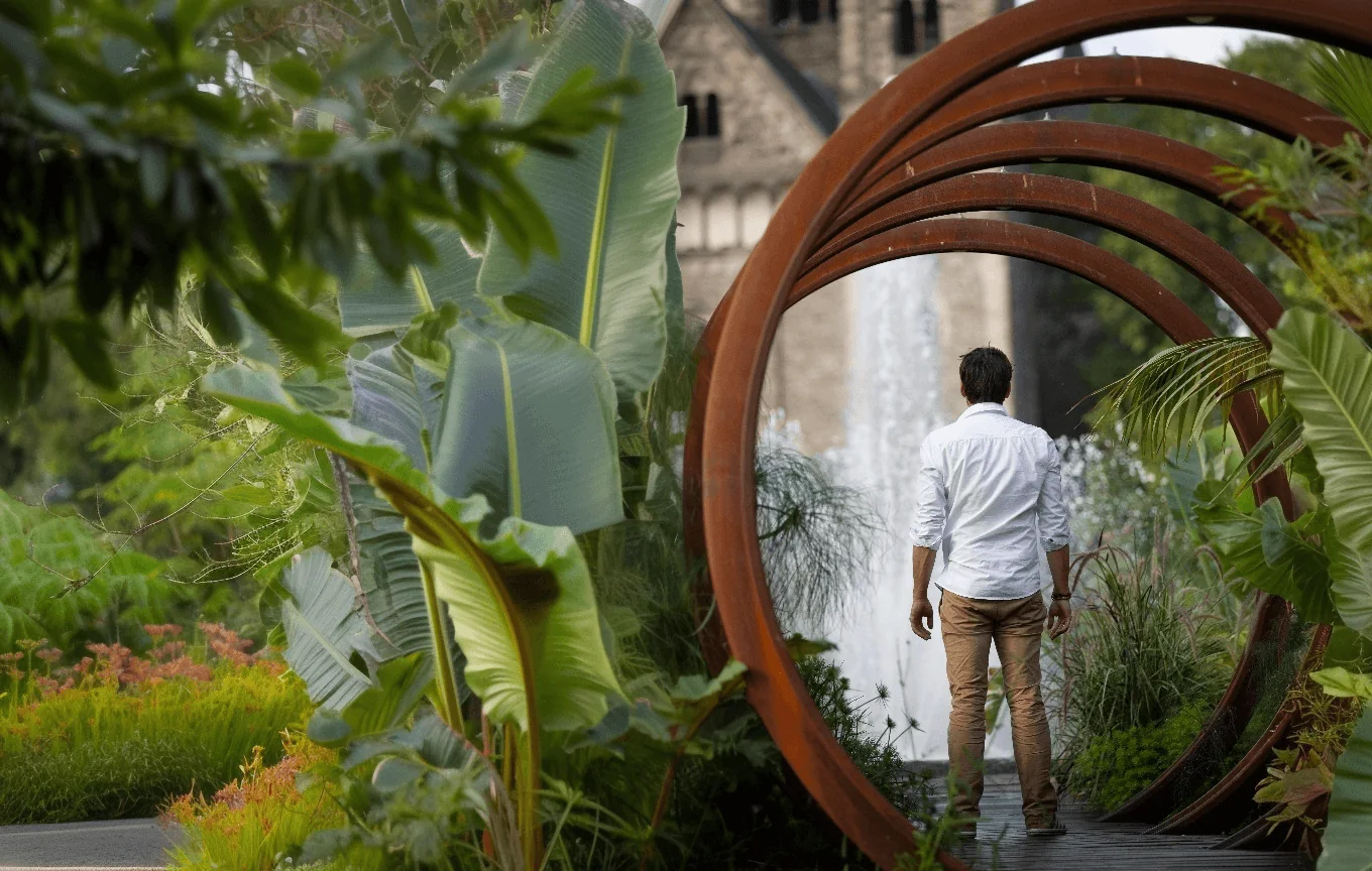 Guillaume, de dos, entre dans un tunnel d'arches rouillées au milieu d'un jardin luxuriant.