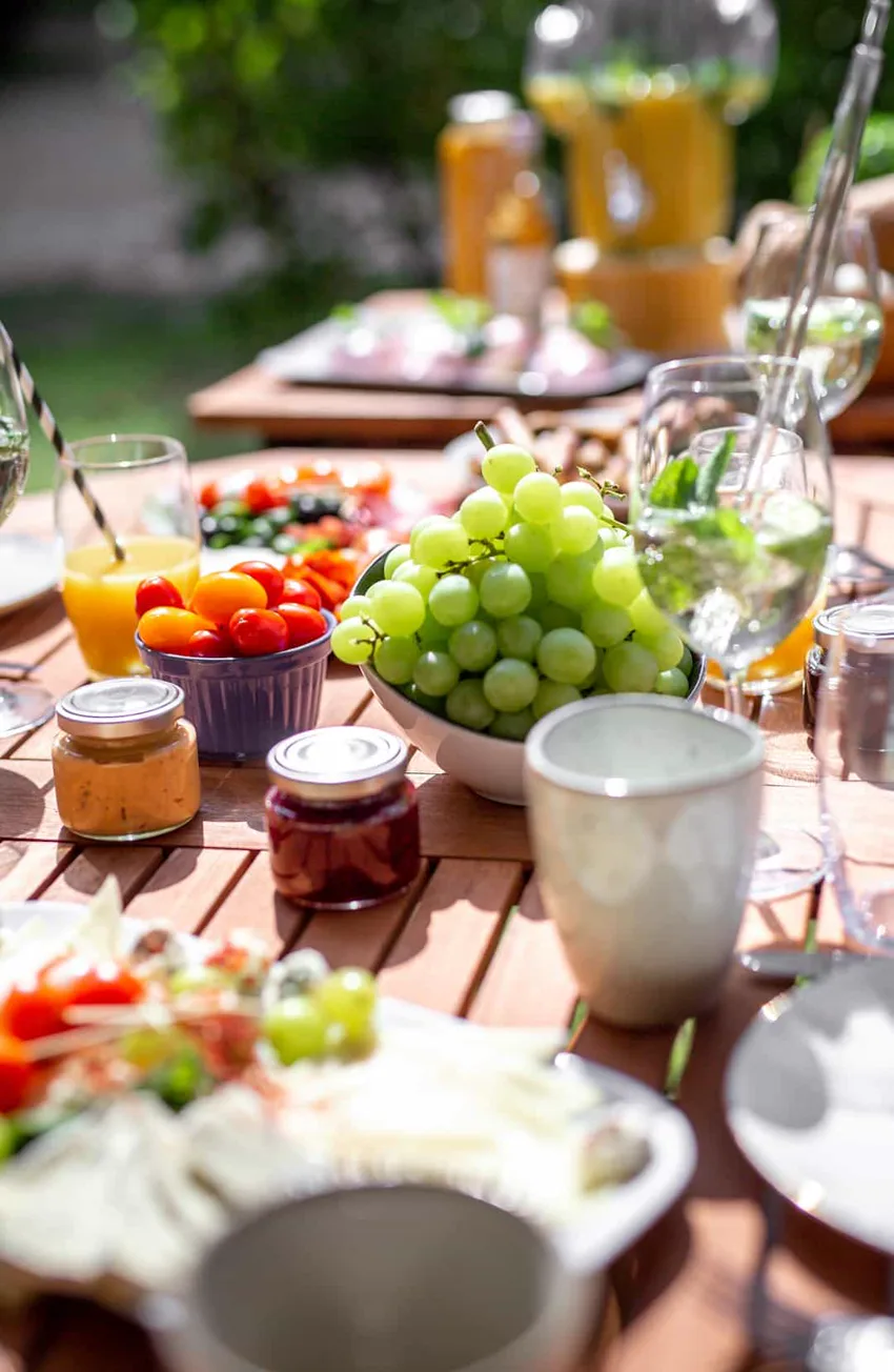 Une table dressée au soleil : raisin, tomates cerises, confitures, jus de fruits et eau à la menthe.